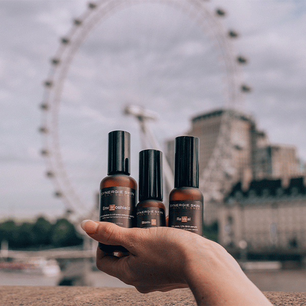 Women's hand holding up Synergie Skin skincare products in front of the London Eye